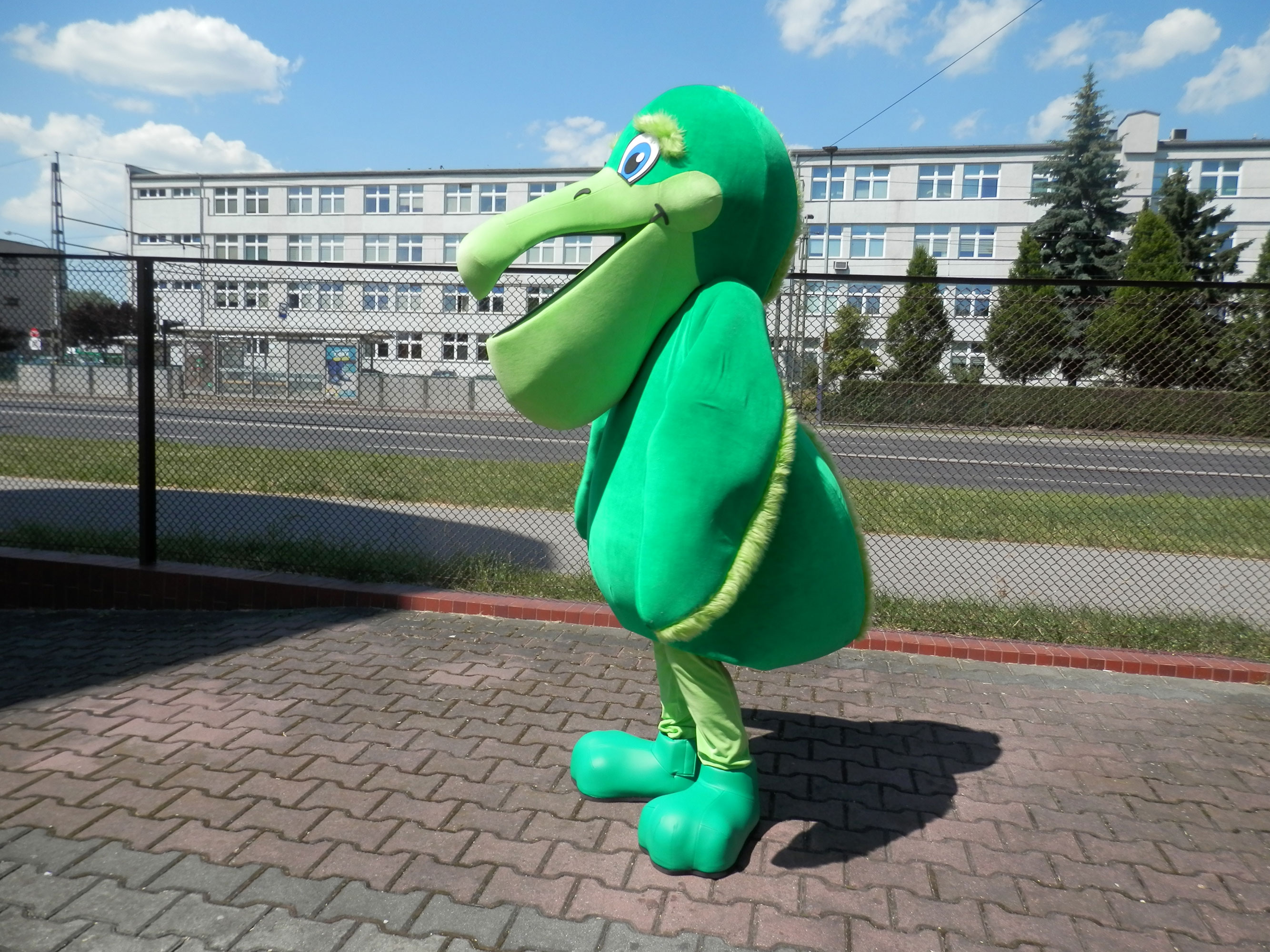 Na fotografii widnieje kostium reklamowy w postaci zielonego pelikana pozujący do zdjęcia obok siedziby firmy Elabika